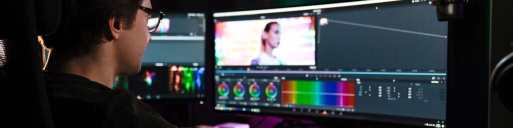 Back view of a young white man video editor sitting in front of a big screen working on a video montage indoors